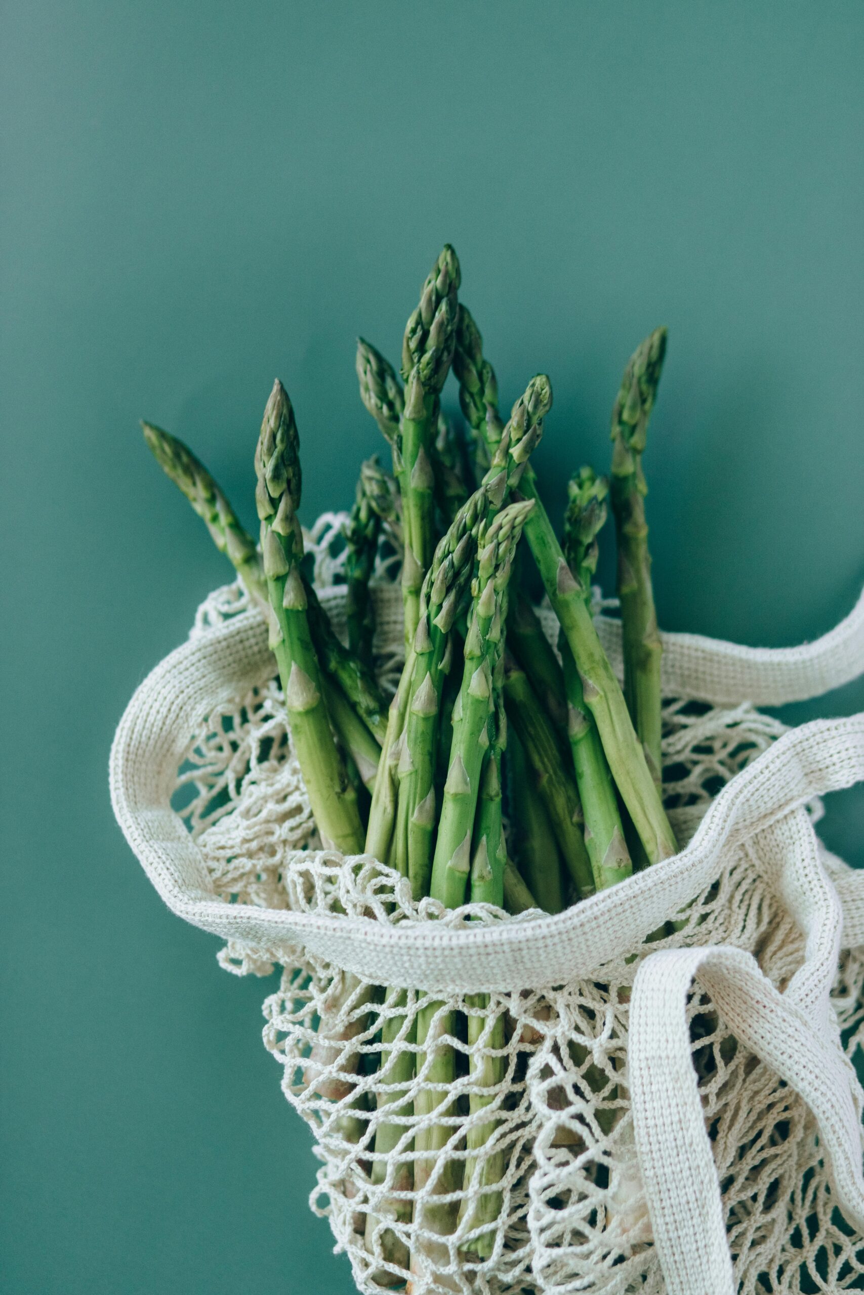 Vertical shot of fresh asparagus in a net bag against a green background, symbolizing healthy eating.